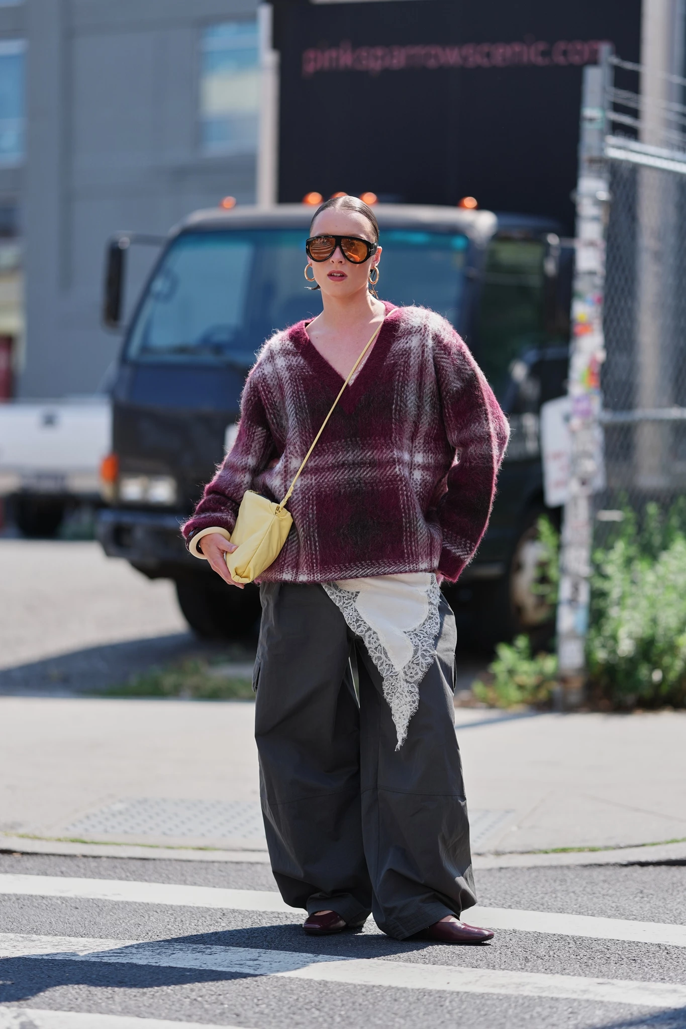 Γυναίκα φοράει πουλόβερ με καρό σε μπορντό και χακί παντελόνι, κρατώντας μια μικρή, κίτρινη τσάντα χιαστί.