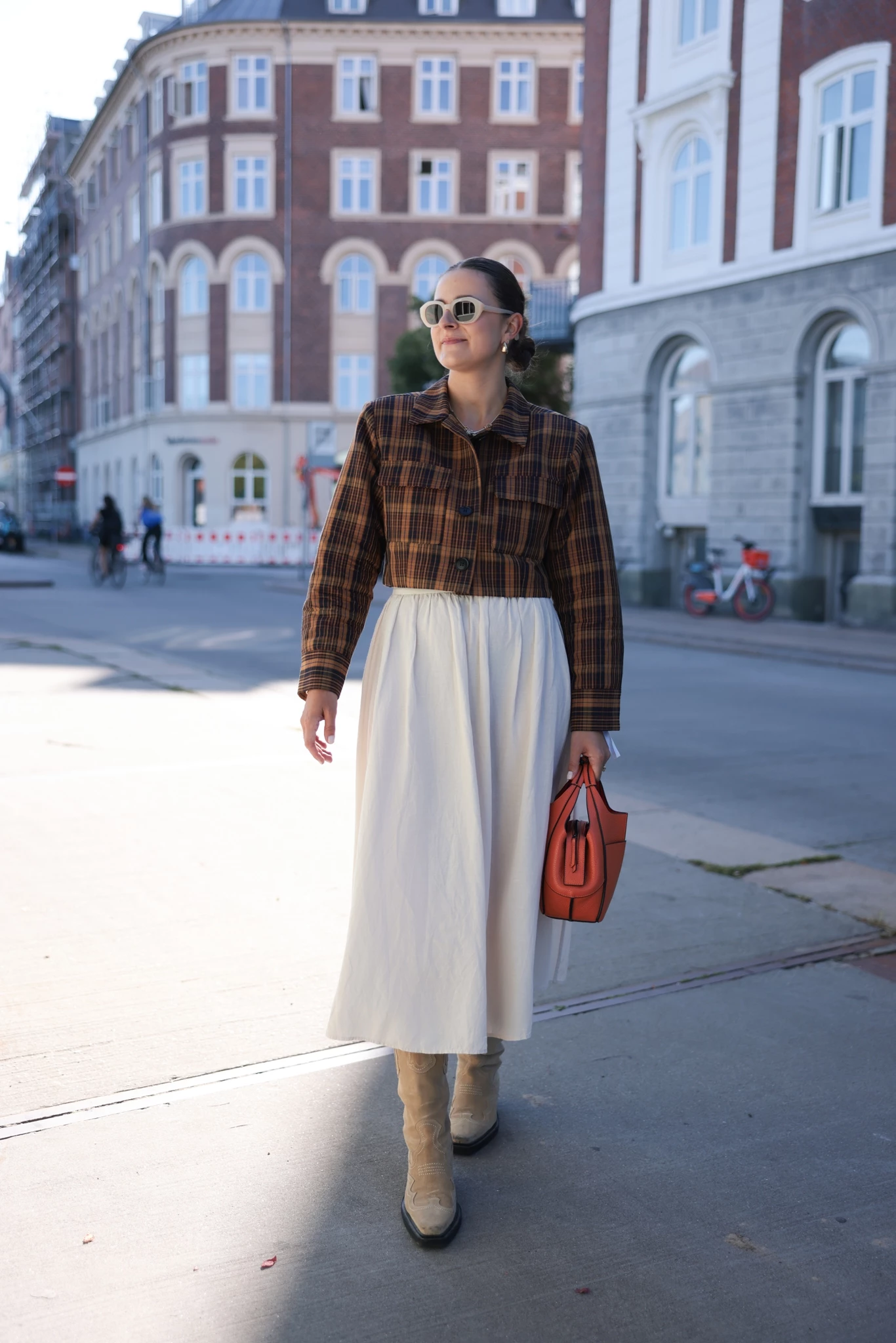 Γυναίκα με λευκή μίντι φούστα, καρό τζάκετ και western μπότες σε μπεζ. Κρατάει κόκκινη τσάντα.