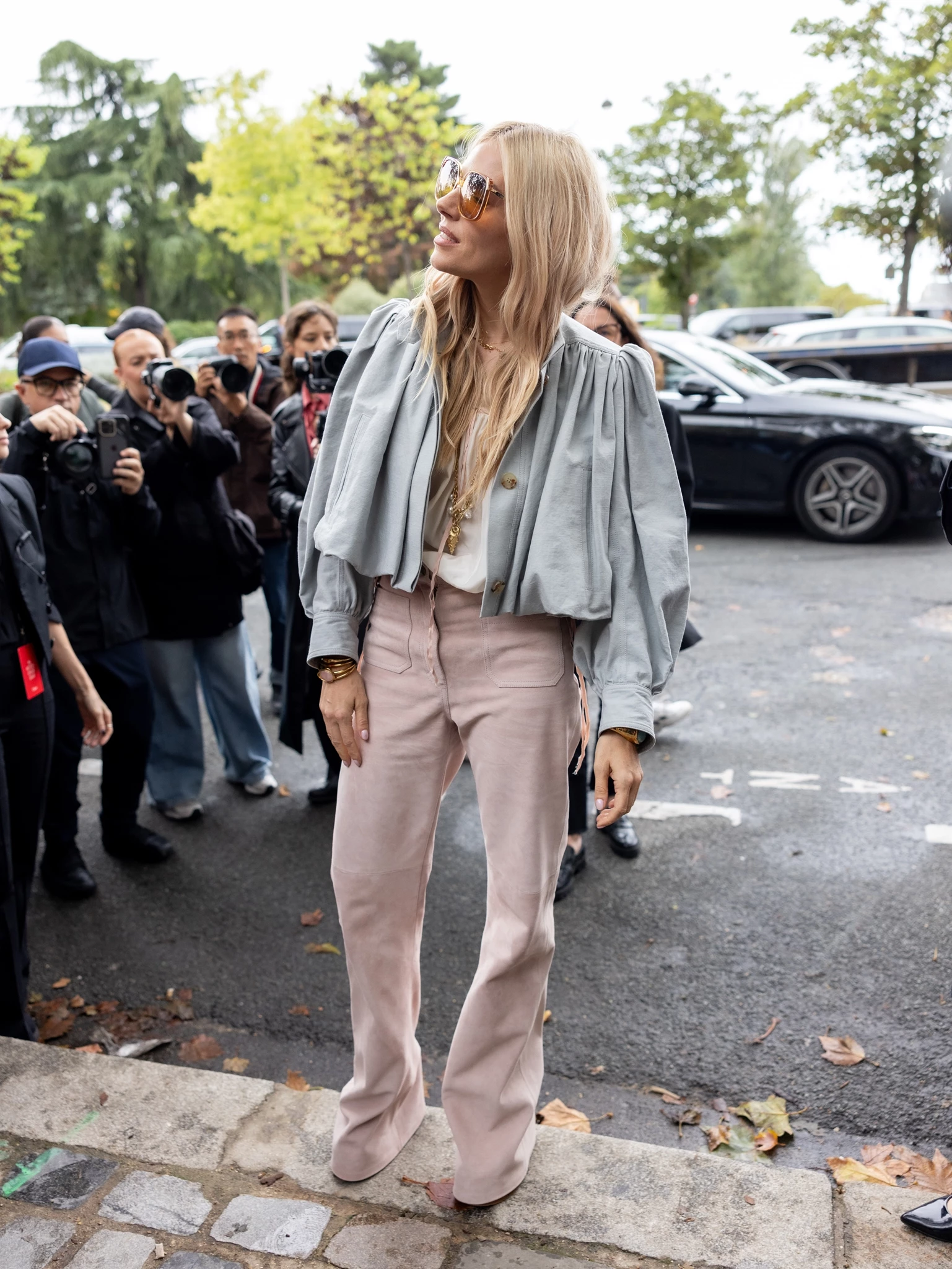 Γυναίκα με ροζ κοτλέ παντελόνι καμπάνα και κοντό γκρι σακάκι με ογκώδη μανίκια (puffy sleeves) ποζάρει μπροστά σε πλήθος φωτογράφων.