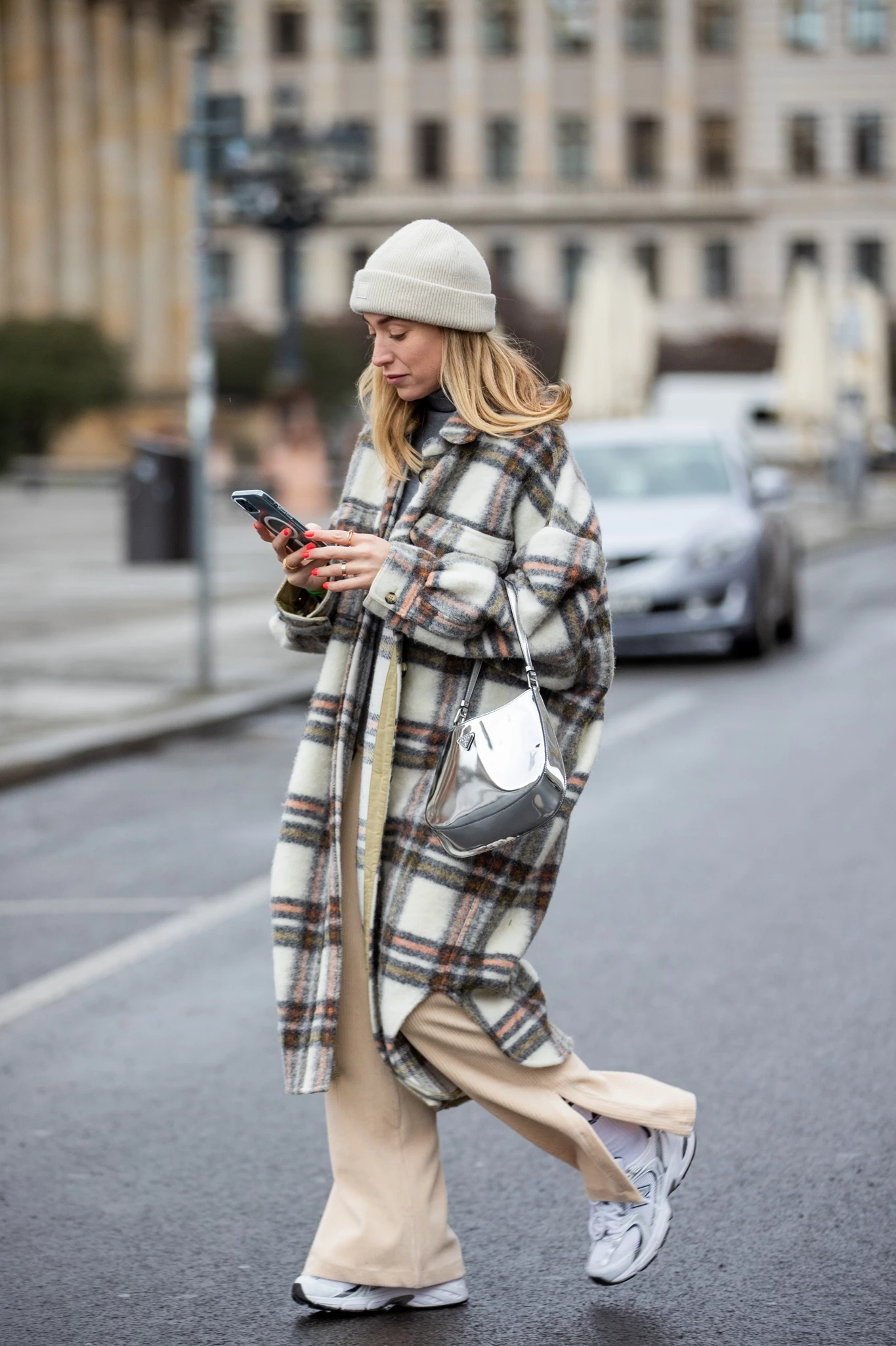 Γυναίκα με μακρύ καρό παλτό, μπεζ φαρδύ κοτλέ παντελόνι, λευκό σκούφο και αθλητικά παπούτσια, περπατά στον δρόμο κοιτώντας το κινητό της.
