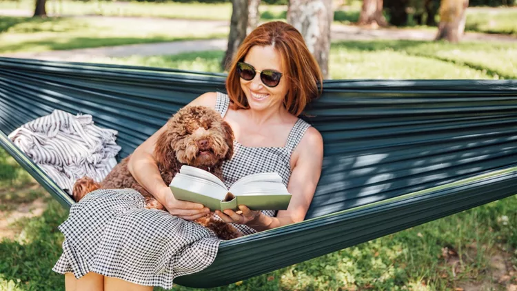Woman reading book relaxing in hammock with her fluffy brown Maltipoo dog on sunny day. Both looking content and happy. This outdoor scene captures joy of bonding with pets and enjoying togetherness