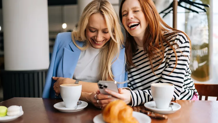 Portrait of two cheerful beautiful women friends sitting in cafe indoors, looking at phone and talking, laughing. Happy pretty girlfriends using social media