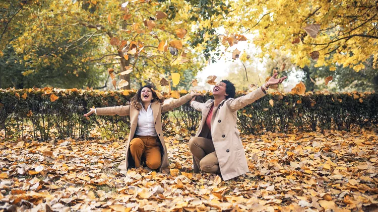 Two smiling young women friends throwing leaves in the park full of fall colors