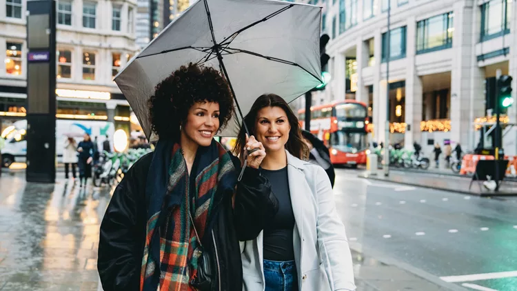 Two smiling young women walking under umbrella in rainy london