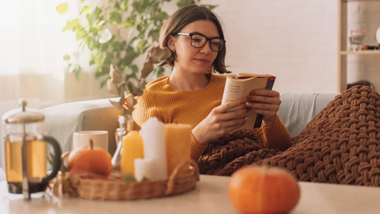 Lady reads a book in blanket on the sofa in front of teapot and candles