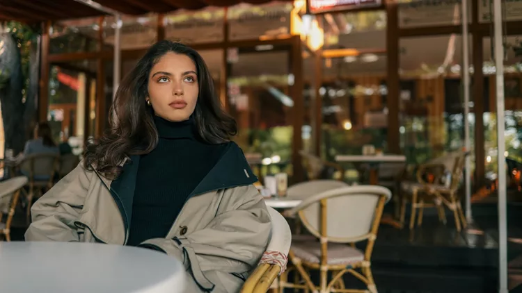 Stylish young woman in a trench coat and black turtleneck sitting alone at a table in a cozy outdoor cafe, looking thoughtful and composed