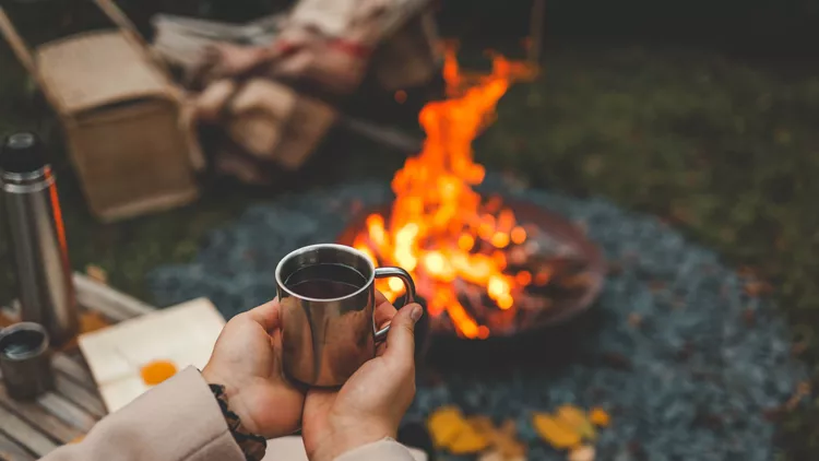 A cozy outdoor scene featuring a person holding a warm drink in a metal mug by a campfire, with a thermos, book, and autumn leaves, peaceful autumn vibes
