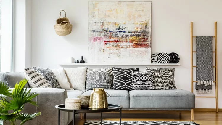 Patterned pillows on grey corner sofa in living room interior with table and painting. Real photo