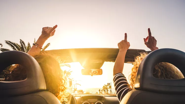 Couple of woman friends traveling and driving having a lot of fun dancing in the car with opened roof and summer vacation sunset ocean in front - concept of friendship together and nice lifestyle for independent girls
