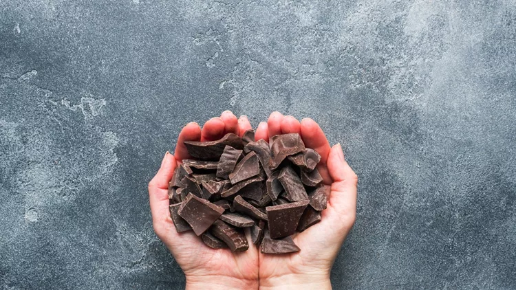 A handful of crushed dark chocolate in women's hands palms on a dark textured background.