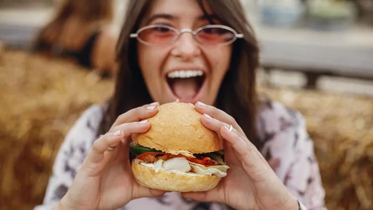 Street food festival. Stylish hipster girl in sunglasses eating delicious vegan burger at street food festival. Happy boho woman tasting and biting burger with vegetables in summer street.