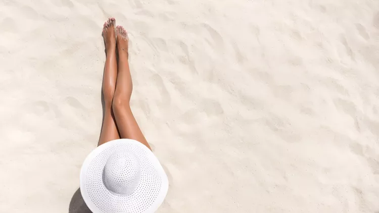 Summer holiday fashion concept - tanning woman wearing sun hat at the beach on a white sand shot from above