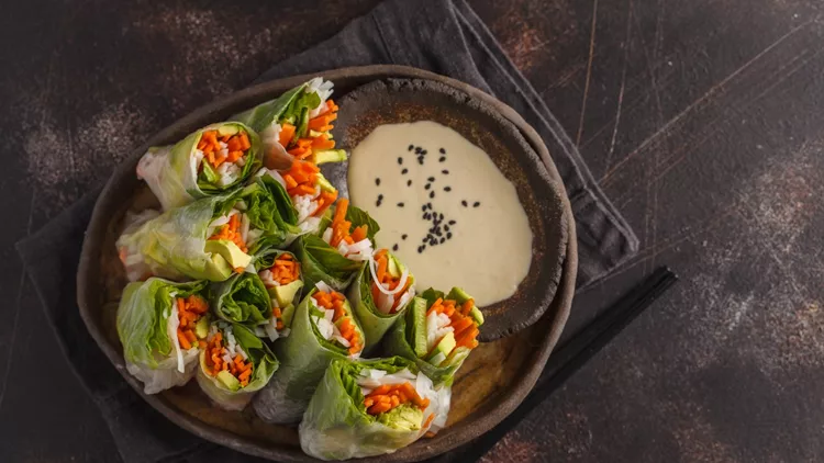 Fresh handmade vegan asian spring rolls with rice noodles, avocado, carrots and tahini dressing on black dish, dark background. Top view, copy space.