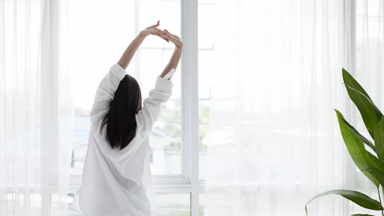 Asian woman waking up in her bed fully rested and open the curtains in the morning to get fresh air