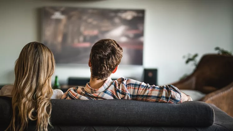 Back view of a couple watching TV on sofa at home.