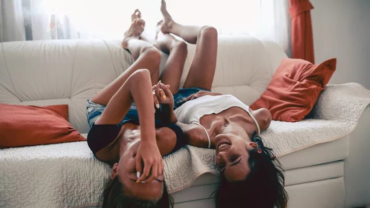 Two Laughing Sensual Females Lying on Sofa and Holding Hands