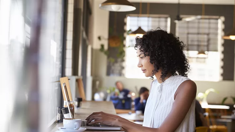 Businesswoman By Window Working On Laptop In Coffee Shop
