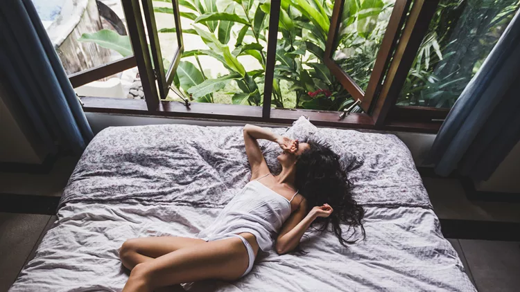 Woman in white lingerie lounging in bed in the morning, view from window on tropical garden. Lifestyle photo