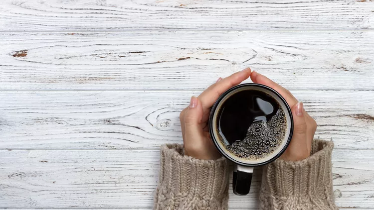 Woman holding cup of hot coffee on rustic wooden table, closeup photo of hands in warm sweater with mug, winter morning concept, top view