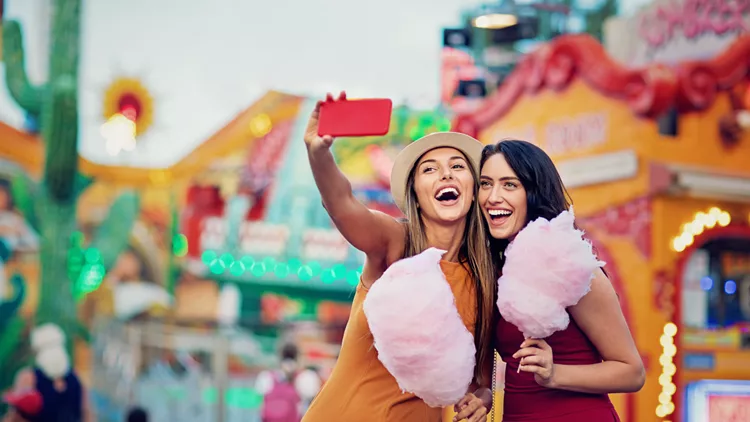 Happy girlfriends are taking selfie/making video call and make fun together in fun fair