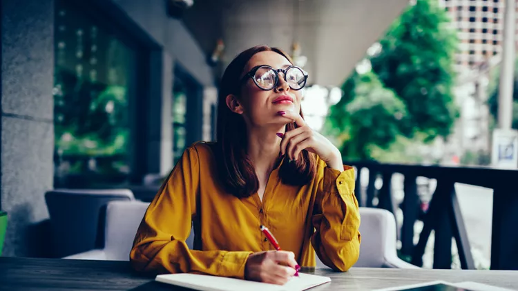 Pondering cute talented student in cool spectacles looking away and thinking while preparing for upcoming exams.Pensive charming redhead hipster girl writing down information to notepad in cafe