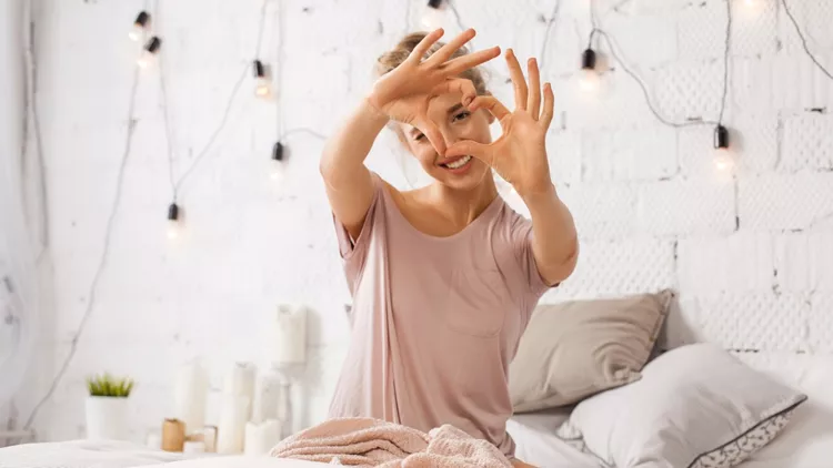 Woman sitting on bed and shows gesture heart with fingers
