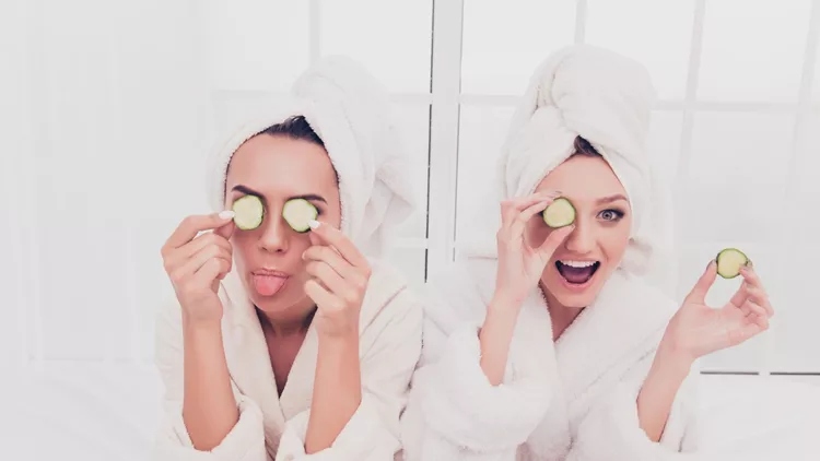 Funny young girls in bathrobes making mask with cucumber and having fun