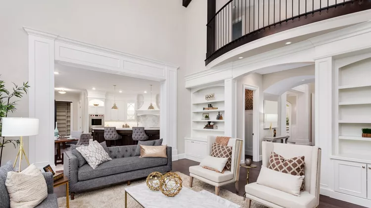 Beautiful living room interior with tall vaulted ceiling, loft area, hardwood floors and fireplace in new luxury home. Has view of kitchen and dining area, and loft.