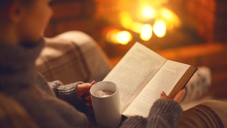 book and cup of coffee in hands of girl on  winter evening near fireplace