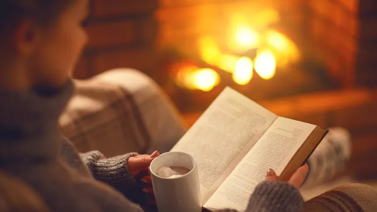 book and cup of coffee in hands of girl on  winter evening near fireplace