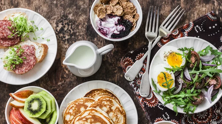 Greek yogurt with whole grain cereals and berry sauce, pancakes, arugula, cherry tomatoes, boiled eggs salad, kiwi, apples fruit, salami and cream cheese sandwiches on a wooden background, top view. Flat lay breakfast table