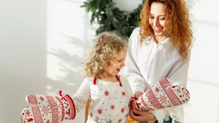 Little hard working child wears kitchen glove and apron, going to help her mother cook dinner, has happy expression. Mother and daughter prepare festive supper on Christmas. Happy family in kitchen