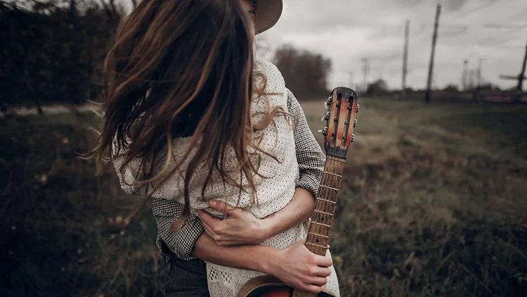 Hipster musician couple hugging in field, handsome man embracing gypsy woman in white dress, guitar closeup