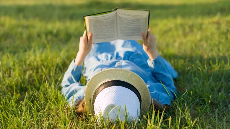 girl-in-dress-and-hat-lies-on-green-grass-reading-book-view-from-picture-id970740862