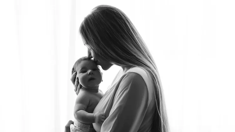 black and white picture of mother kissing little baby boy in front of curtains at home