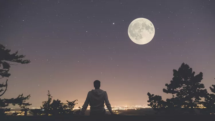 Man enjoying the view from hill above city. Full moon and stars on the sky.