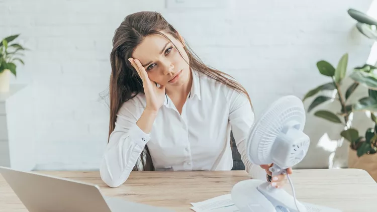 upset businesswoman sitting at workplace with paperwork, laptop and electric fan