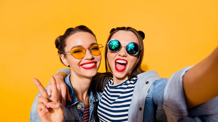 Two glad positive grinning lady stand in glasses spectacles street style stylish trendy cool casual denim jeans clothes isolated on yellow background in take picture on cellular make hollywood smile