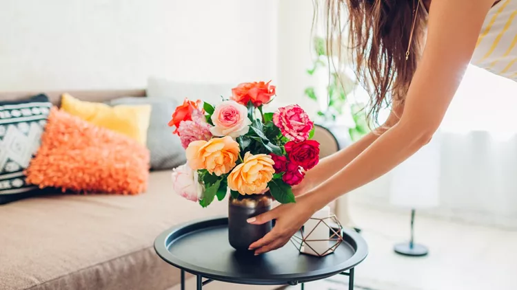Woman puts vase with flowers roses on table. Housewife taking care of coziness in apartment. Interior and decor