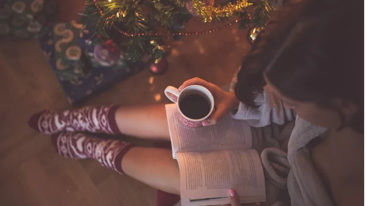 girl-by-christmas-tree-with-coffee-cup-reading-a-book-picture-id614639662