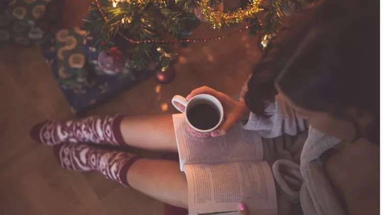 girl-by-christmas-tree-with-coffee-cup-reading-a-book-picture-id614639662