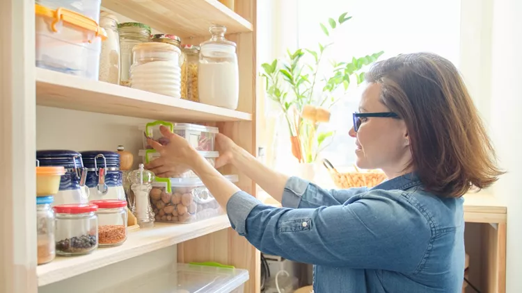 Interior of wooden pantry with products for cooking. Adult woman taking kitchenware and food from the shelves