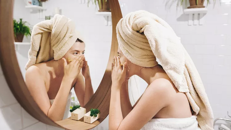 Young happy woman in towel applying organic face scrub and looking at round mirror in stylish bathroom. Girl making facial massage, peeling and cleaning skin on face. Skin Care and Hygiene