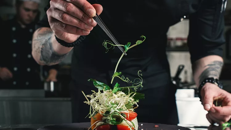 Chef finishing healthy salad on a black plate with tweezers. almost ready to serve it on a table