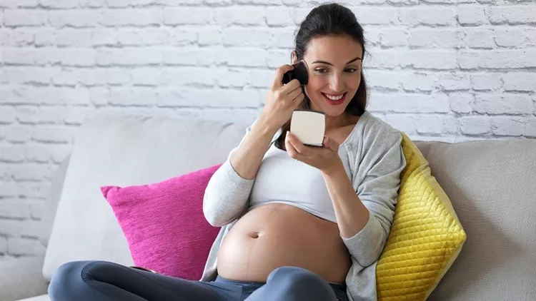 Beautiful pregnant young woman doing her makeup while sitting on sofa at home.