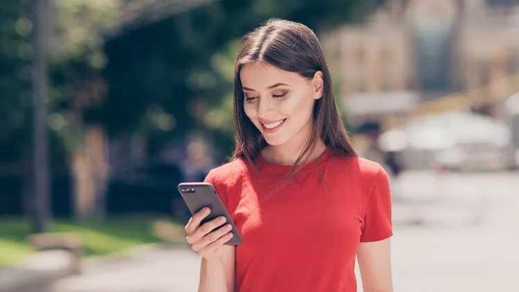 Fit girl smiling and looking into the phone holding her hand in the pocket of blue jeans on a hot summer day