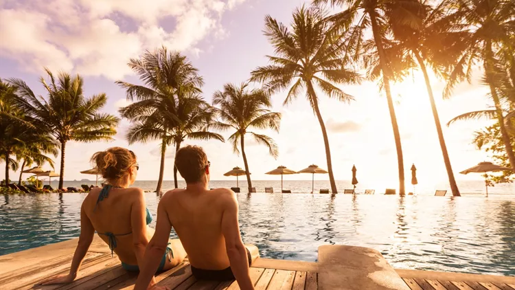 Couple enjoying beach vacation holidays at tropical resort with swimming pool and coconut palm trees near the coast with beautiful landscape at sunset, honeymoon destination