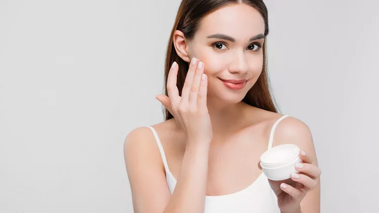 beautiful girl applying cosmetic cream on face, isolated on grey