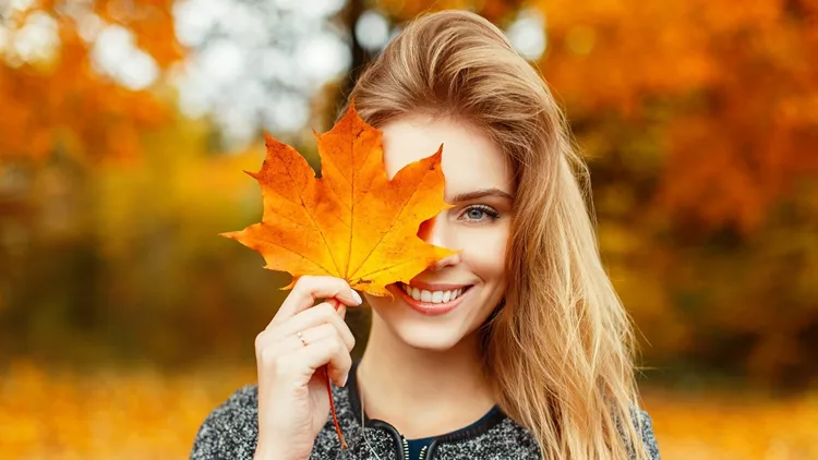 Pretty young happy young woman in stylish clothes with a beautiful smile holds in her hand an autumn gold-yellow leaf near face on a warm autumn day in the park. Funny happy girl enjoys weekend.