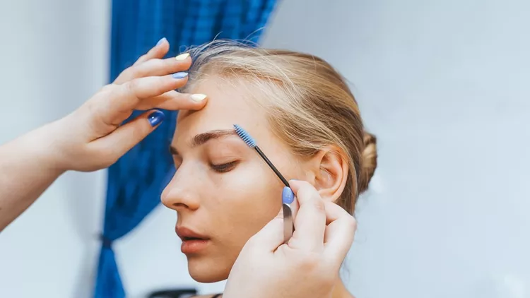 A female cosmetologist performs eyebrow correction on a beautiful model in a beauty salon.